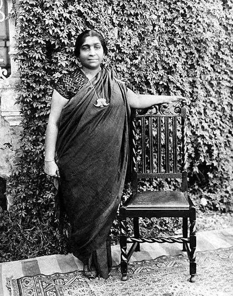 Full-length photograph of Sarojini Naidu standing beside a carved wooden chair in front of an ivy-covered wall, wearing a dark sari and a pendant.