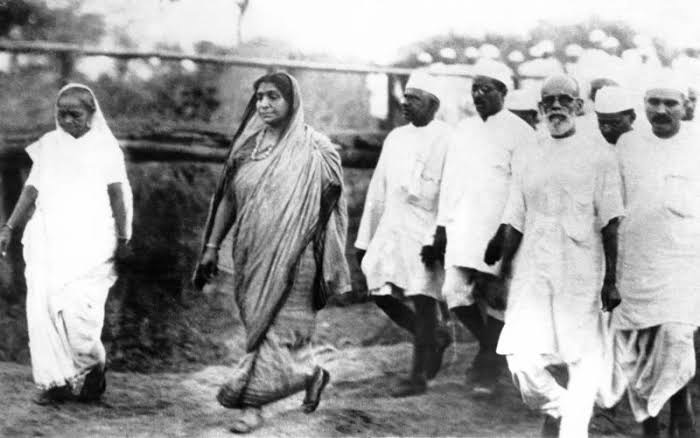 Sarojini Naidu walking in a group of marchers on an open road, two women at the front and a line of men in white khadi caps behind.