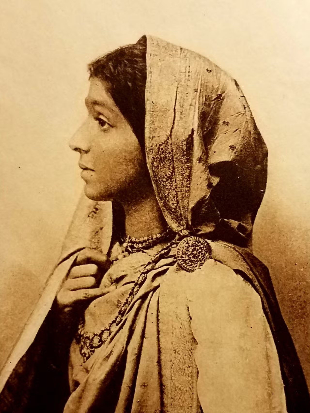 Sepia studio portrait of a young Sarojini Naidu in profile, wearing an embroidered sari drawn over her hair and a fine chain necklace.