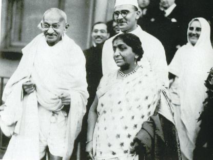 Gandhi in a white shawl standing beside Sarojini Naidu in a light sari, with other members of a delegation behind them, photographed in London.